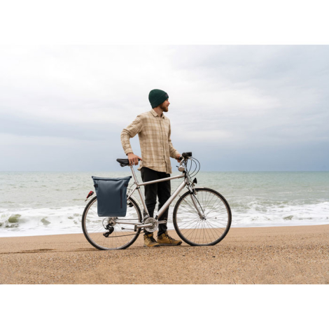 Un homme portant un bonnet vert se tient à côté d'un vélo sur une plage, face à la mer. Un sac à dos bleu foncé est fixé à l'arr