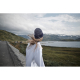 Une femme vue de dos porte un bonnet bleu foncé et un haut blanc, face à un paysage de montagnes et de lac.