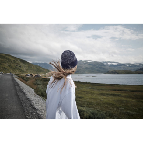 Une femme vue de dos porte un bonnet bleu foncé et un haut blanc, face à un paysage de montagnes et de lac.