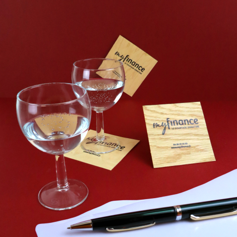 Plusieurs sous-verres carrés en bois clair, avec logo et textes noirs, disposés autour de deux verres à pied sur fond rouge. Vei