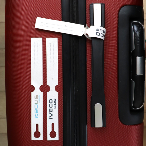 Deux étiquettes de bagage blanches avec logos IVECO BUS et KEOLIS, posées sur une valise rouge. Une troisième est attachée à la 