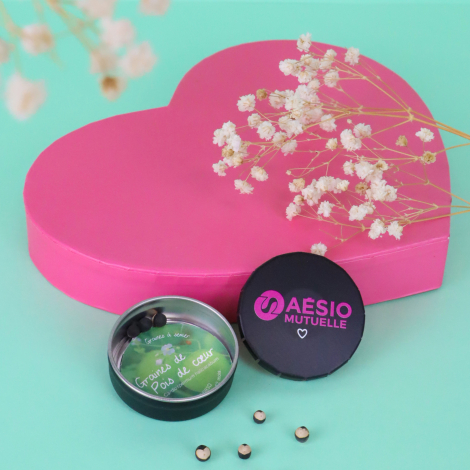 Boîte ronde noire personnalisée AESIO Mutuelle, ouverte avec des graines de pois de cœur, devant une grande boîte rose en forme 