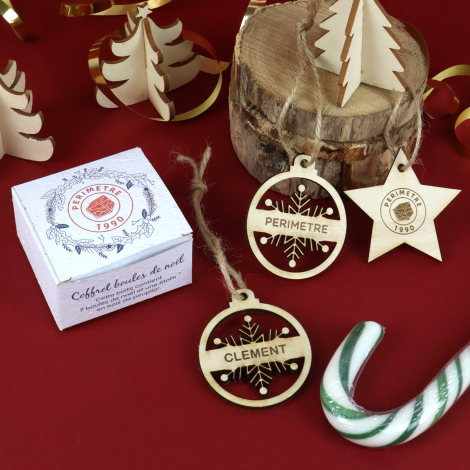 Gros plan sur un coffret de Noël personnalisé et trois suspensions en bois gravé, deux boules ajourées et une étoile, entourés d