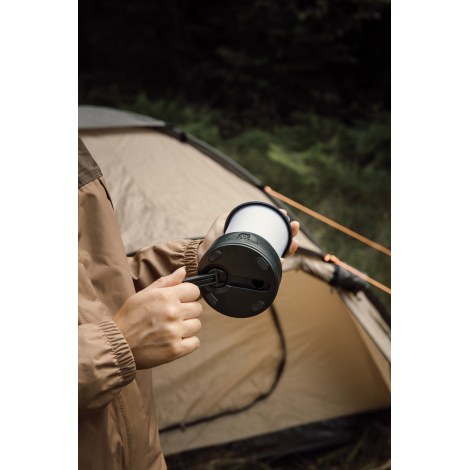 Une personne en veste marron actionne la manivelle située sous la lanterne, tenue horizontalement devant une tente de camping.