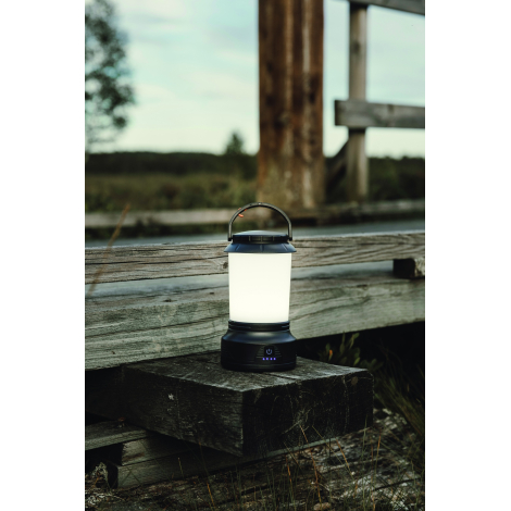 Lanterne noire allumée posée sur une poutre en bois, à proximité d'une barrière rustique dans un environnement naturel.