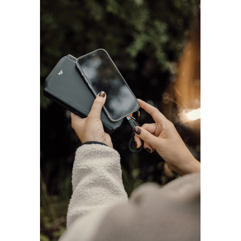 Une femme en veste claire tient un smartphone contre un chargeur noir pliable et approche le connecteur du câble de charge tress