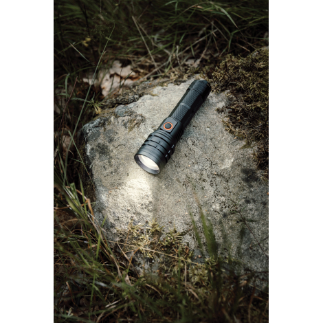 Lampe torche noire allumée, posée sur un rocher couvert de mousse en pleine nature, projetant un faisceau lumineux blanc sur la 