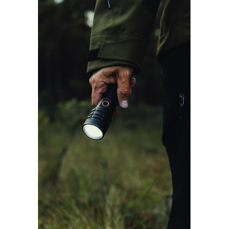 Vue rapprochée d'une lampe torche noire allumée tenue par un homme en veste kaki, avec tête large rainurée et bouton latéral cer