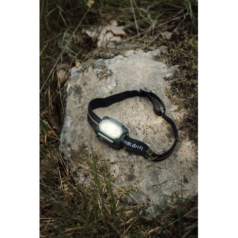 Lampe frontale noire allumée, posée sur un rocher entouré d'herbes et de mousse. Son bandeau élastique est disposé en boucle, av