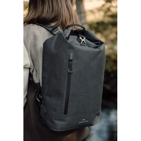 Femme photographiée de dos en extérieur, portant un sac à dos noir à fermeture enroulable, avec poche zippée verticale et bretel