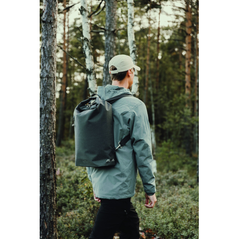 Homme photographié de dos dans une forêt, portant un sac à dos noir à fermeture enroulable, avec bretelles noires et petit motif