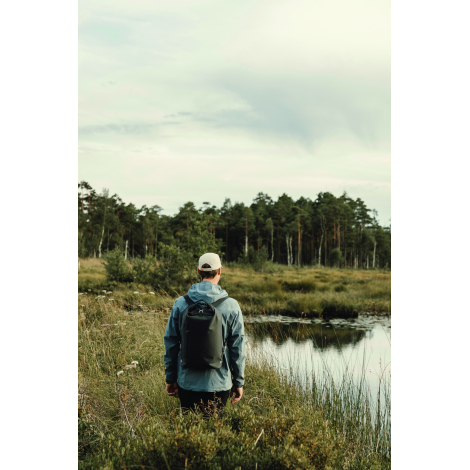 Homme vu de dos, portant un sac à dos noir en marchant au bord d’un étang entouré de végétation et d’arbres.