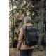 Femme vue de trois quarts dos dans un environnement boisé, portant un sac à dos noir à rabat fermé par une boucle centrale, avec Femme vue de trois quarts dos dans un environnement boisé, portant un sac à dos noir à rabat fermé par une boucle centrale, avec