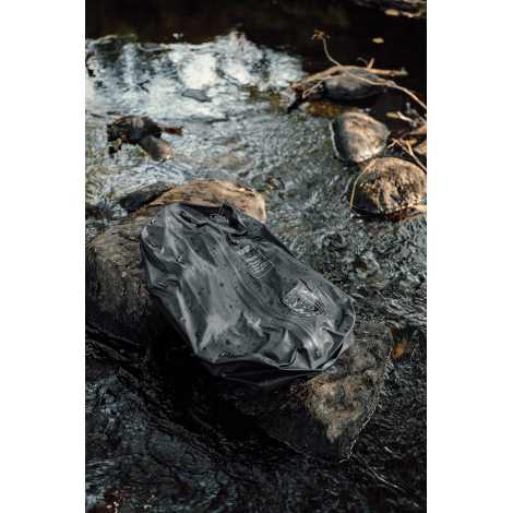 Sac de compression noir posé sur des rochers au bord d'un ruisseau, avec des gouttes d'eau sur sa surface, une fermeture zippée 