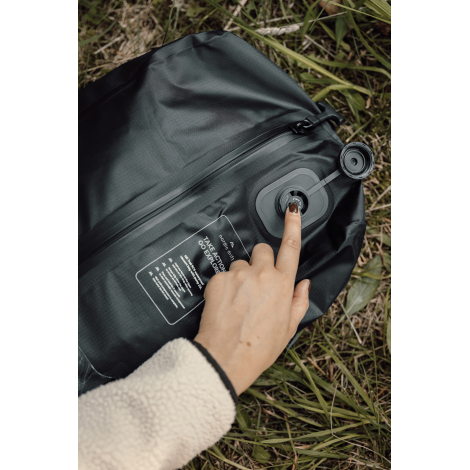 Gros plan sur la valve ronde ouverte du sac de compression noir posé dans l'herbe, avec une main appuyant sur son mécanisme et u