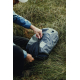 Un homme vêtu d'une veste bleue manipule la fermeture zippée du sac de compression noir posé dans l'herbe, avec valve ronde et p
