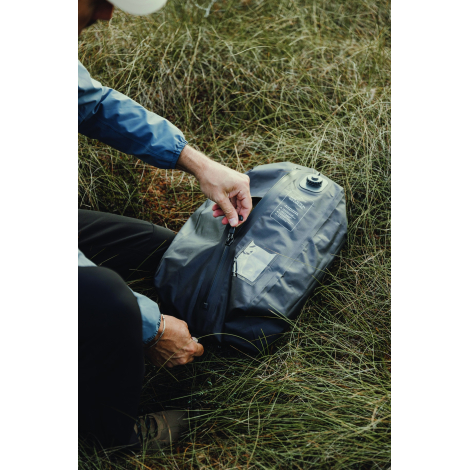 Un homme vêtu d'une veste bleue manipule la fermeture zippée du sac de compression noir posé dans l'herbe, avec valve ronde et p