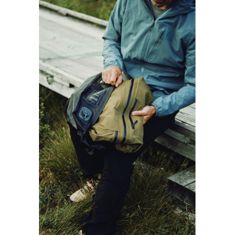Un homme assis sur une structure en bois place un sac beige à fermetures zippées dans le sac de compression noir ouvert, dont la
