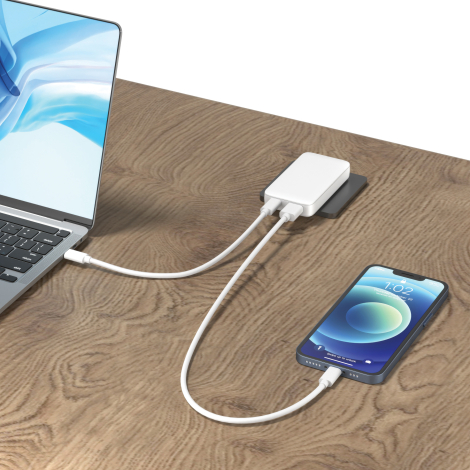 Sur une table en bois, le chargeur plat blanc est relié par deux câbles à un ordinateur portable ouvert et à un smartphone posé 