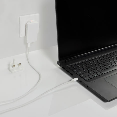 Chargeur blanc branché sur une prise murale, relié par un câble à un ordinateur portable noir ouvert sur un bureau blanc. Une fi