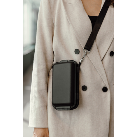 Femme portant un étui de voyage rigide noir en bandoulière sur un manteau beige, illustrant son format compact et son port près 