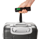Une main soulève une valise rigide grise par sa poignée à l'aide de la sangle noire de la balance à bagages. L'appareil est tenu Une main soulève une valise rigide grise par sa poignée à l'aide de la sangle noire de la balance à bagages. L'appareil est tenu