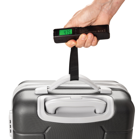 Une main soulève une valise rigide grise par sa poignée à l'aide de la sangle noire de la balance à bagages. L'appareil est tenu