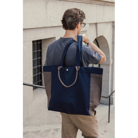 Homme portant à l'épaule un grand sac shopping bleu marine et marron, avec ses longues anses et ses poignées courtes visibles su
