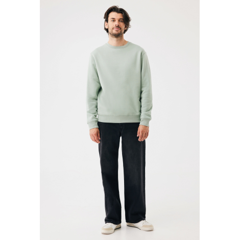 Un homme debout face à l'objectif porte un sweat uni vert sauge à col rond et manches longues, associé à un pantalon noir ample 