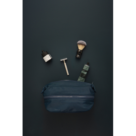 Trousse de toilette bleu marine posée sur un fond sombre, entourée d'accessoires de rasage comprenant un blaireau, un rasoir, un