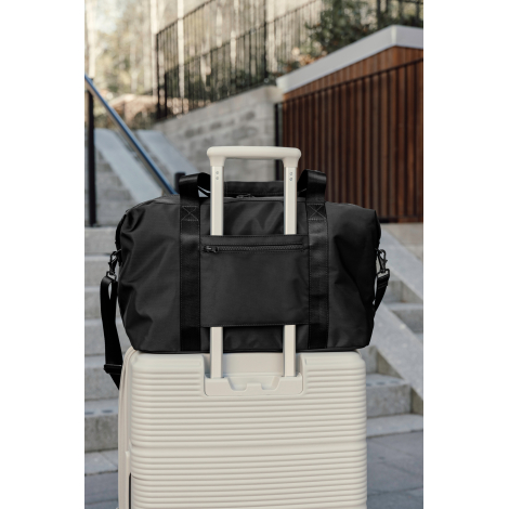 Vue arrière d'un sac de week-end noir fixé sur la poignée télescopique d'une valise rigide blanche, avec une poche zippée horizo