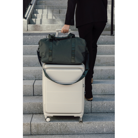 Une personne vêtue de noir tient une valise rigide claire surmontée d'un sac de week-end vert foncé, devant un escalier en pierr
