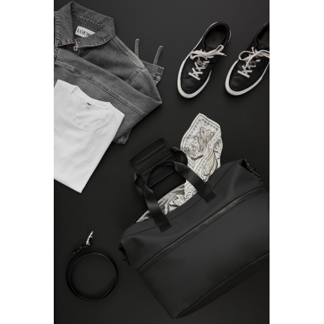 Sac de week-end noir ouvert, vu de dessus sur un fond sombre, contenant des vêtements clairs et un foulard à motifs. Un jean gri