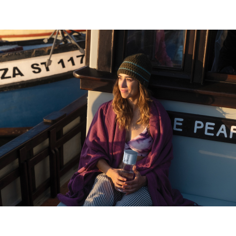 Une femme assise sur un bateau, vêtue d'un bonnet rayé et enveloppée dans une couverture violette, tient une gourde transparente