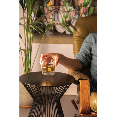 Personne assise dans un fauteuil en cuir marron, tenant un verre de whisky ambré contenant des glaçons métalliques, au-dessus d'