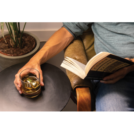 Homme assis dans un fauteuil en cuir marron, un livre ouvert à la main, tenant un verre de whisky ambré avec des glaçons métalli