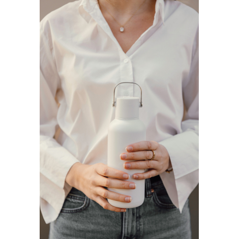 Vue rapprochée d'une femme en chemise blanche et jean gris, tenant à deux mains une bouteille isotherme blanche mate, avec un bo