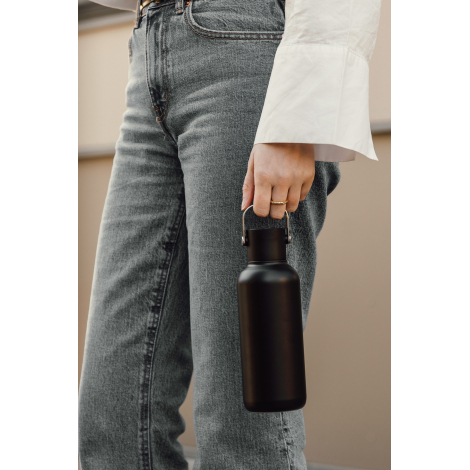 Gros plan sur une femme en jean gris et chemise blanche, portant une bouteille isotherme noire mate par son anse métallique arge