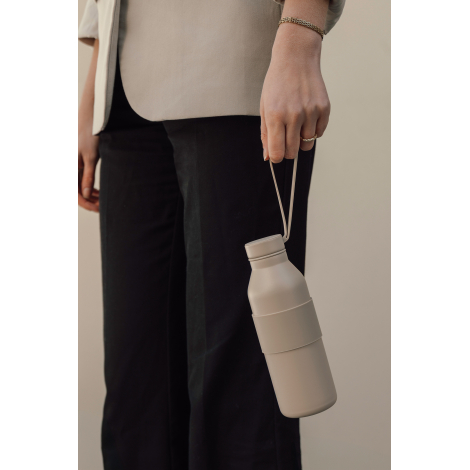 Gros plan sur une femme en blazer beige et pantalon noir, portant une bouteille isotherme beige clair par sa dragonne souple. Le