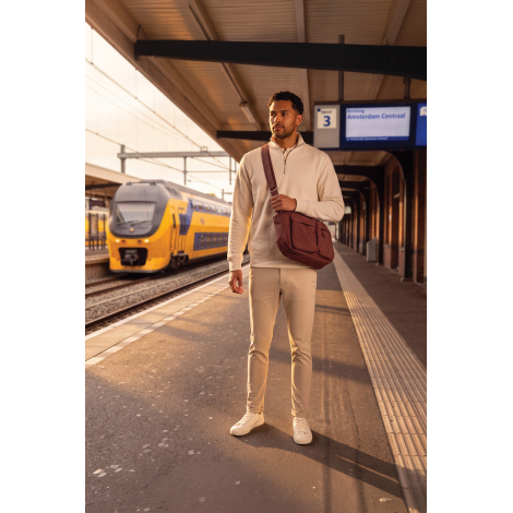 Un homme vêtu d'un ensemble beige et de baskets blanches porte un sac bandoulière brun en travers du buste, avec deux poches ava