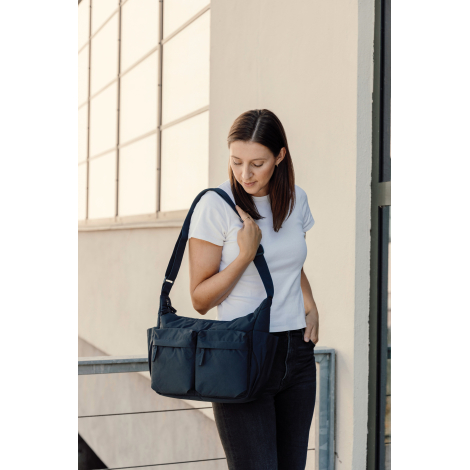 Une femme en t-shirt blanc et pantalon noir porte un sac bandoulière bleu marine à deux poches avant et poches latérales, tout e