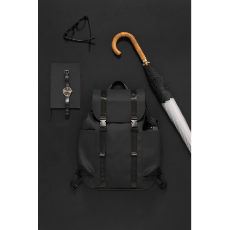Sac à dos noir mat posé à plat sur un fond sombre, entouré d'un carnet avec montre, de lunettes et d'un parapluie à manche en bo