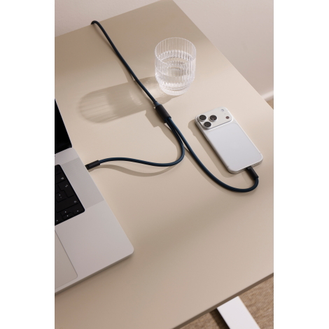 Câble multifonction bleu foncé reliant un ordinateur portable argenté à un smartphone blanc, posé sur une table beige. Un verre 