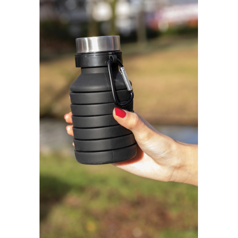 Bouteille pliable noire en silicone compactée, tenue dans une main en extérieur. Le corps présente plusieurs anneaux rapprochés,