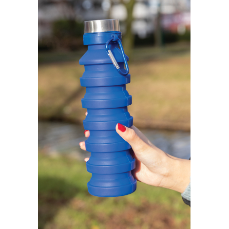 Bouteille pliable bleue en silicone entièrement déployée, tenue dans une main en extérieur. Son corps à larges anneaux superposé