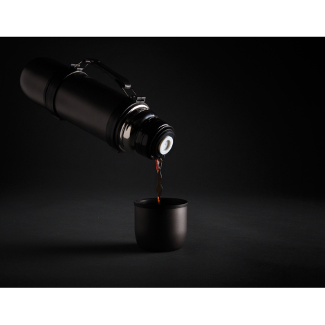 Sur fond noir, une bouteille isotherme gris anthracite inclinée verse un liquide brun dans une tasse assortie. Le bouchon verseu