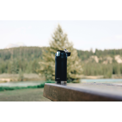 Mug isotherme noir mat posé sur une table extérieure en pierre, avec bouchon à clapet ouvert et base argentée. À l'arrière-plan 