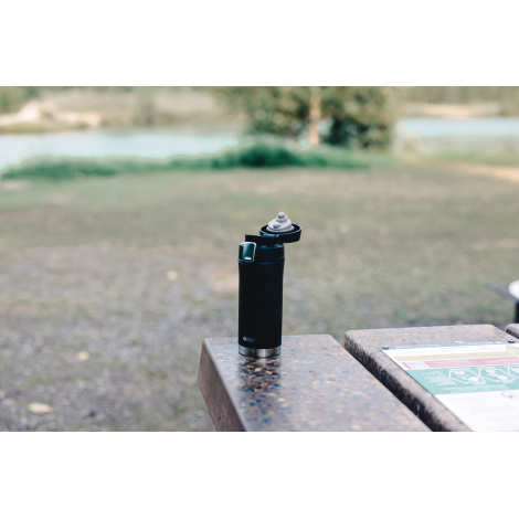 Mug isotherme noir mat avec bouchon ouvert, posé sur une table en pierre à côté d'un panneau d'information. La vue légèrement pl