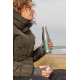 Femme aux cheveux bouclés, vêtue d'une veste kaki, assise près de la mer. Elle tient une bouteille isotherme argentée ouverte, o
